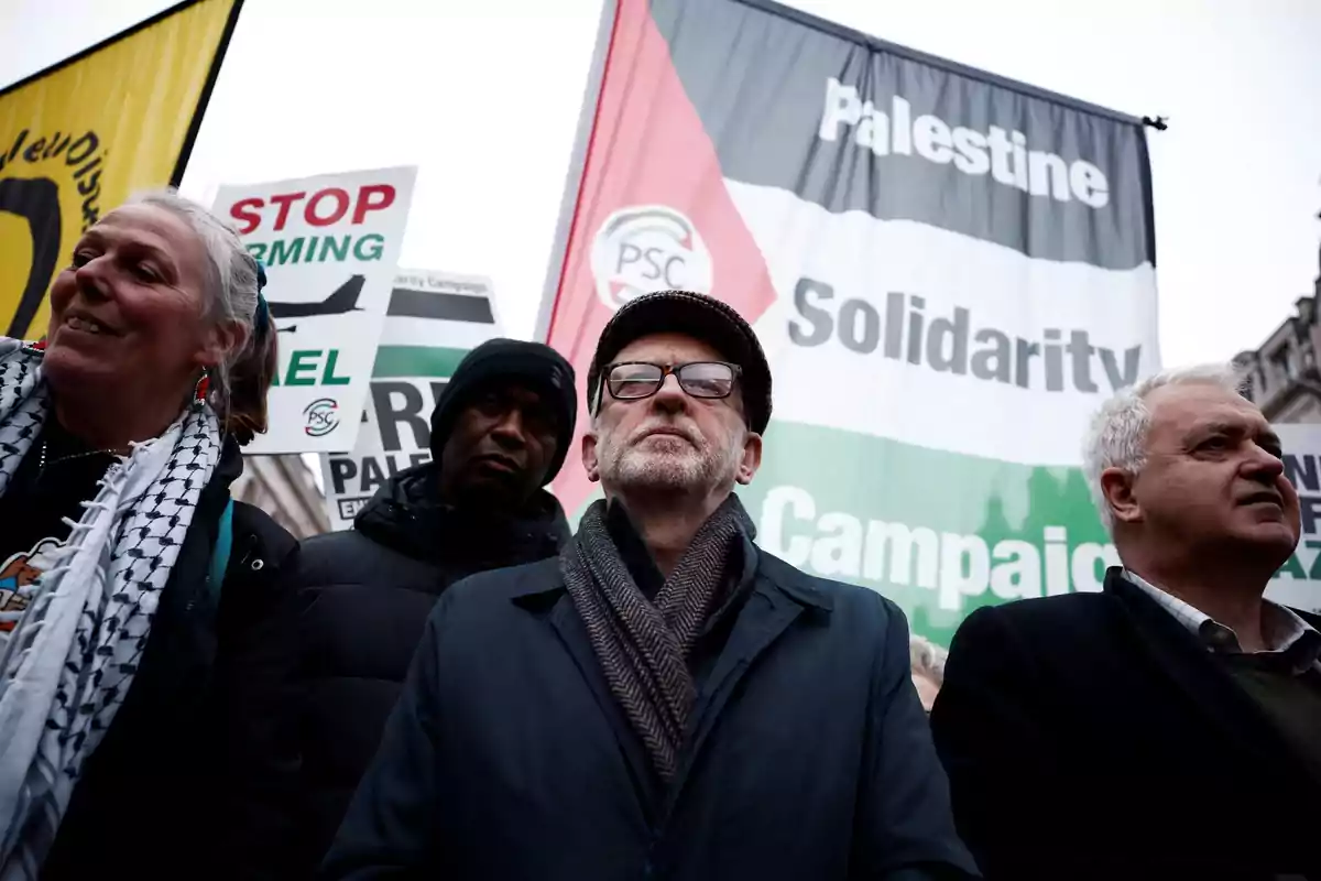 Varias personas participan en una manifestación con pancartas y una gran bandera que dice Palestine Solidarity Campaign