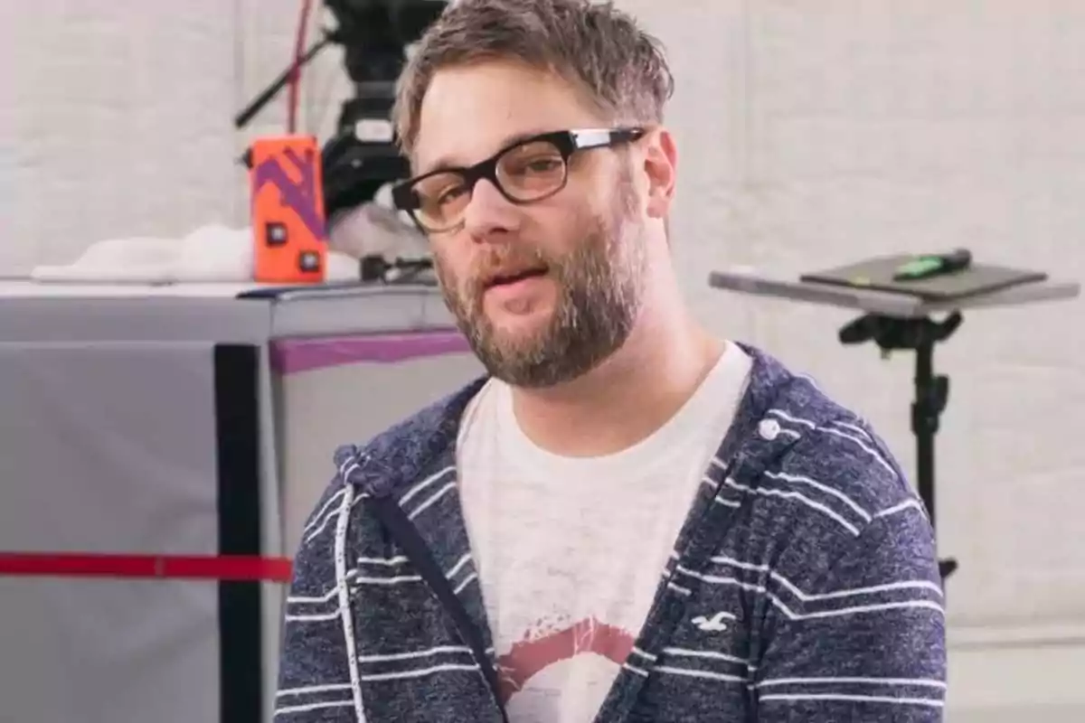 Hombre con barba y gafas de pasta negra vistiendo una sudadera azul con rayas blancas y camiseta blanca en un entorno interior con equipo de grabación al fondo Hombre con barba y gafas de pasta negra vistiendo una sudadera azul con rayas blancas y camiseta blanca en un entorno interior con equipo de grabación al fondo