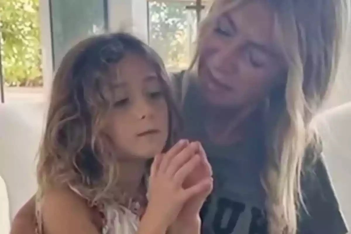 Una mujer rubia abraza a una niña de cabello rizado mientras ambas miran algo entre las manos de la niña en una habitación iluminada por la luz natural que entra por la ventana. Una mujer rubia abraza a una niña de cabello rizado mientras ambas miran algo entre las manos de la niña en una habitación iluminada por la luz natural que entra por la ventana.