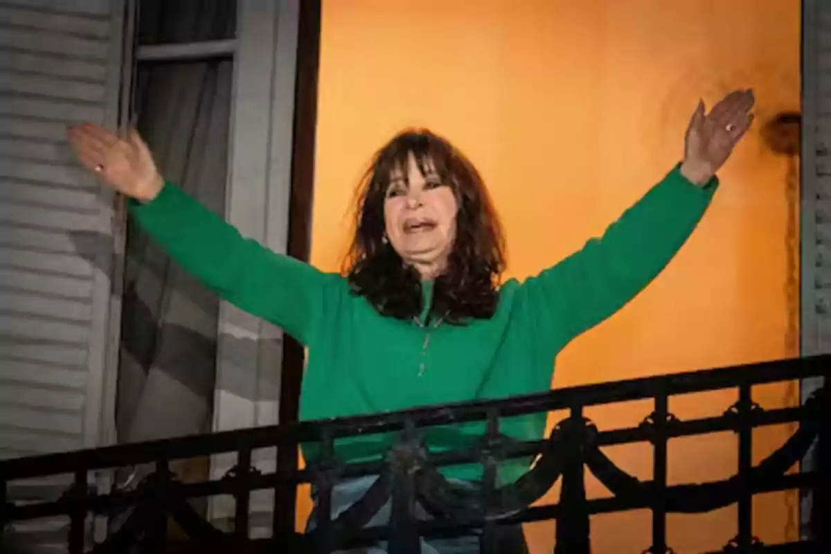 Dark-haired woman in a green sweater greets with open arms from a balcony in front of an orange-lit wall