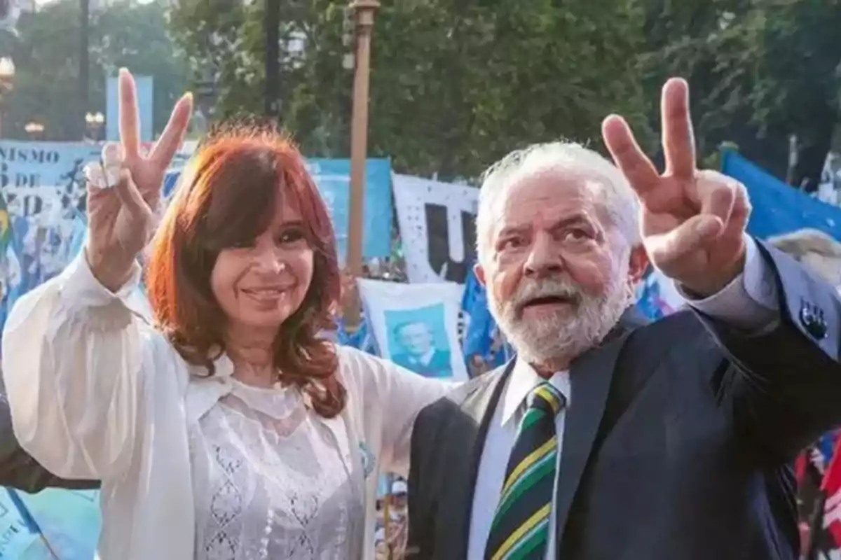 Dos personas sonríen y hacen el gesto de la victoria con los dedos mientras están rodeadas de banderas y pancartas en un evento al aire libre Dos personas sonríen y hacen el gesto de la victoria con los dedos mientras están rodeadas de banderas y pancartas en un evento al aire libre