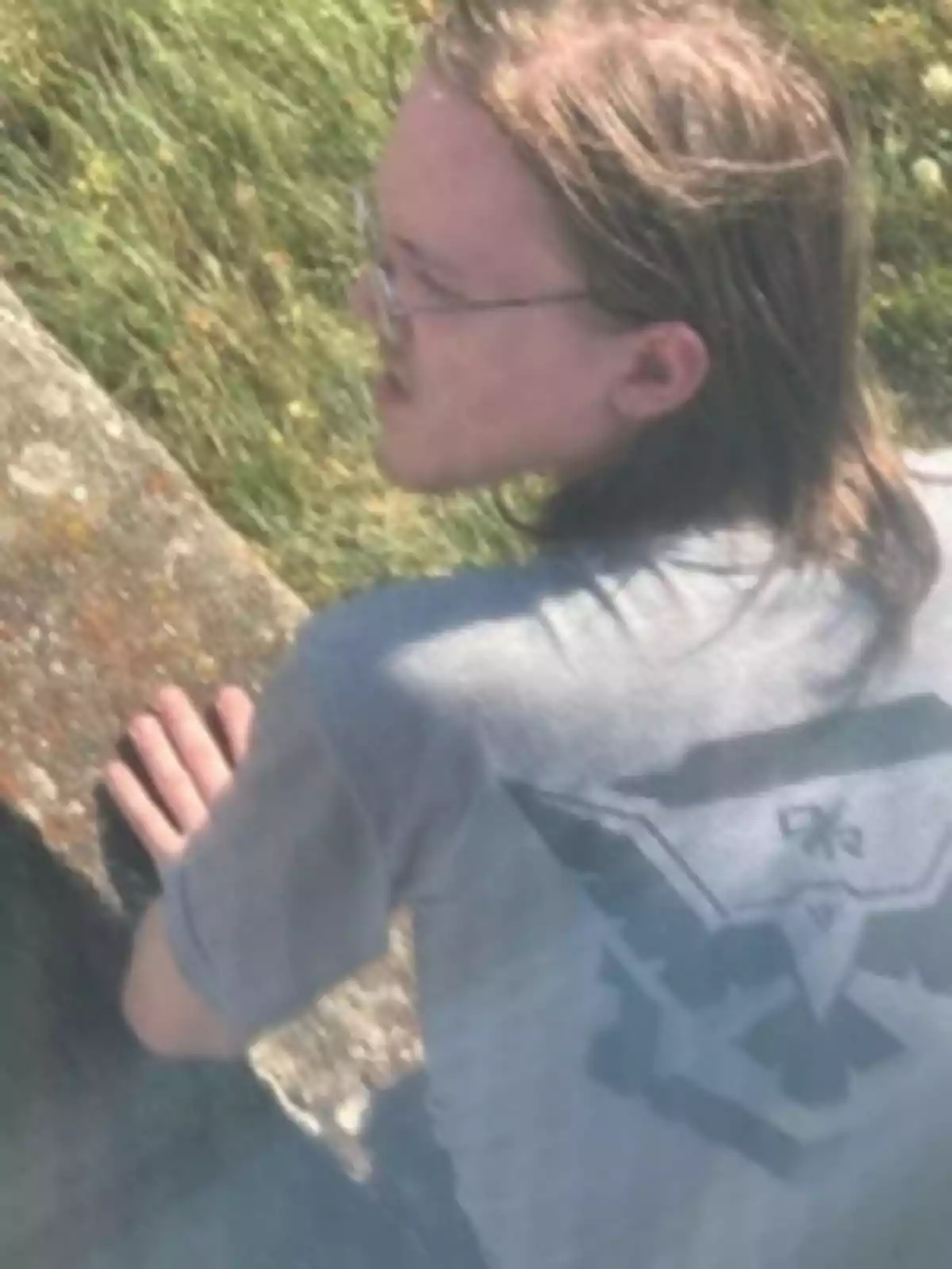 Person with long hair and glasses sitting outdoors, resting their hands on a concrete surface and wearing a gray t-jersey with an eagle design on the back. Person with long hair and glasses sitting outdoors, resting their hands on a concrete surface and wearing a gray t-jersey with an eagle design on the back.