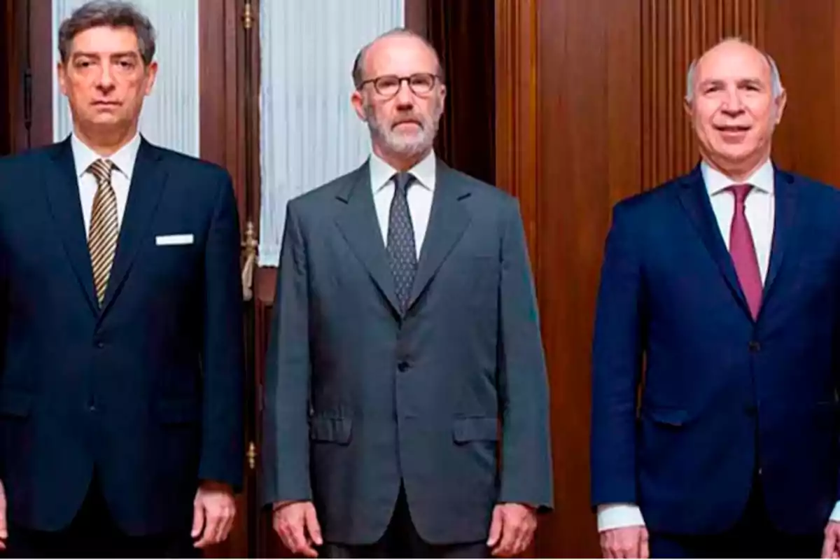 Tres hombres de traje posan juntos en un ambiente formal con fondo de madera y cortinas blancas Tres hombres de traje posan juntos en un ambiente formal con fondo de madera y cortinas blancas
