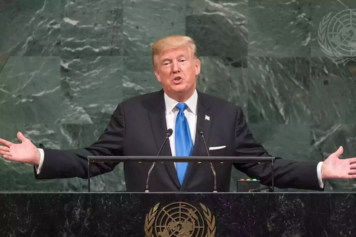 Blond-haired man in a dark suit and blue tie speaking at a podium with the UN emblem in the background
