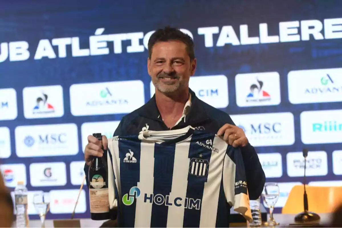 Hombre sonriente sostiene una camiseta de fútbol del club Talleres y una botella durante una conferencia de prensa con fondo de logotipos y el nombre del club