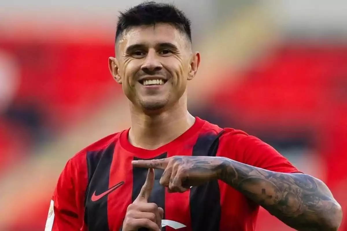 Futbolista sonriente con uniforme rojo y negro celebrando con las manos en un estadio desenfocado.