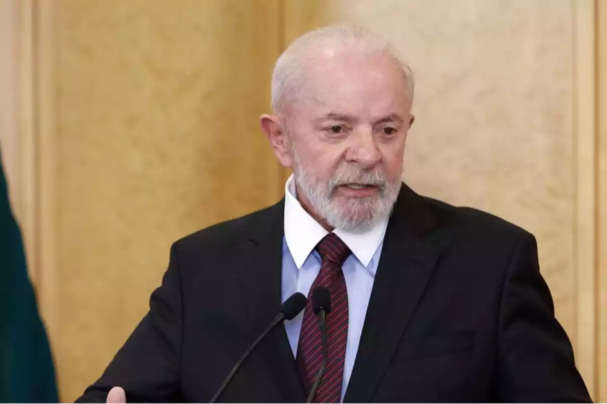 Older man with gray hair and beard wearing a dark suit and red tie speaking in front of two microphones Older man with gray hair and beard wearing a dark suit and red tie speaking in front of two microphones