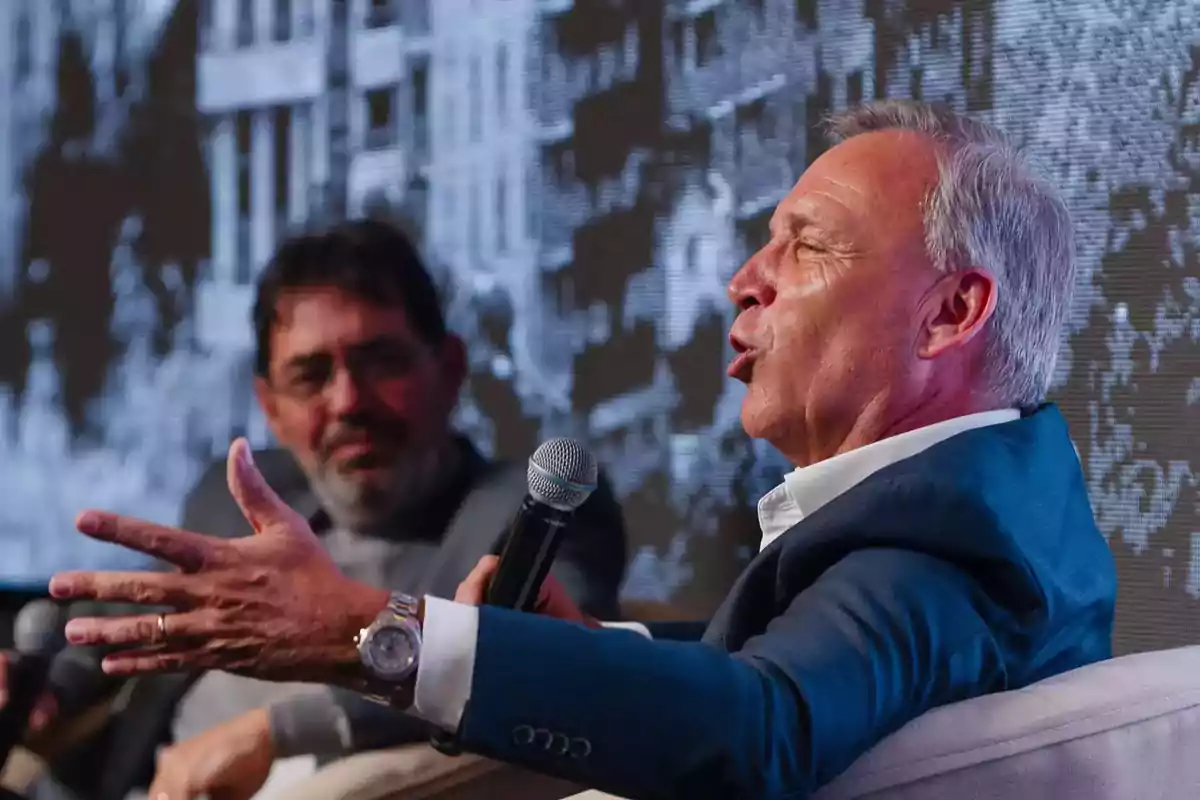 Dos hombres sentados conversan en un evento mientras sostienen micrófonos uno de ellos gesticula con la mano y al fondo se observa una pantalla con una imagen en blanco y negro Dos hombres sentados conversan en un evento mientras sostienen micrófonos uno de ellos gesticula con la mano y al fondo se observa una pantalla con una imagen en blanco y negro