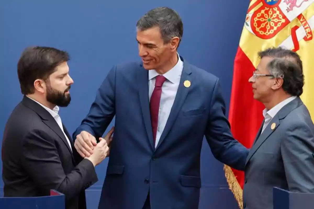 Tres hombres con traje conversan frente a una pared azul y una bandera de España Tres hombres con traje conversan frente a una pared azul y una bandera de España