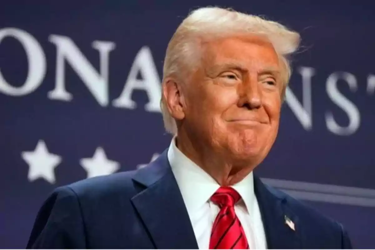 Hombre de cabello rubio con traje azul y corbata roja sonriendo frente a un fondo azul con letras blancas y estrellas