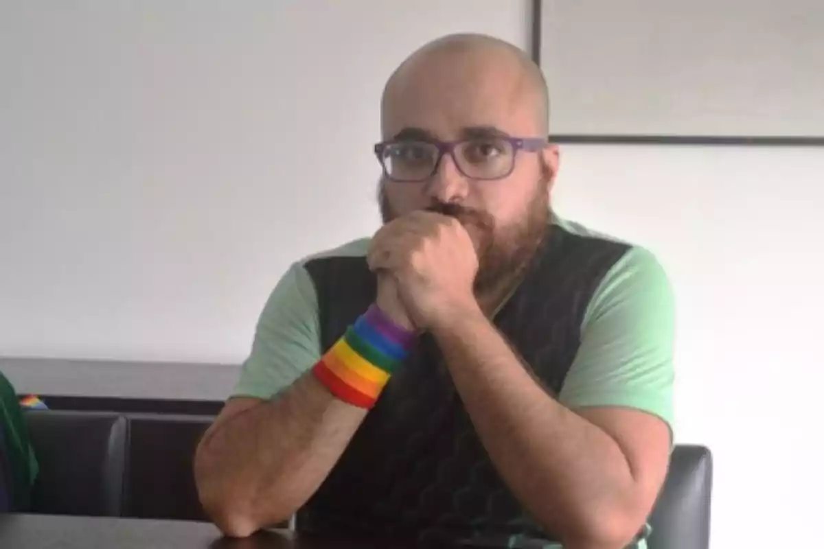 Person with glasses and a beard sitting at a table, wearing a green t-jersey and a bracelet with the colors of the rainbow on their left wrist.