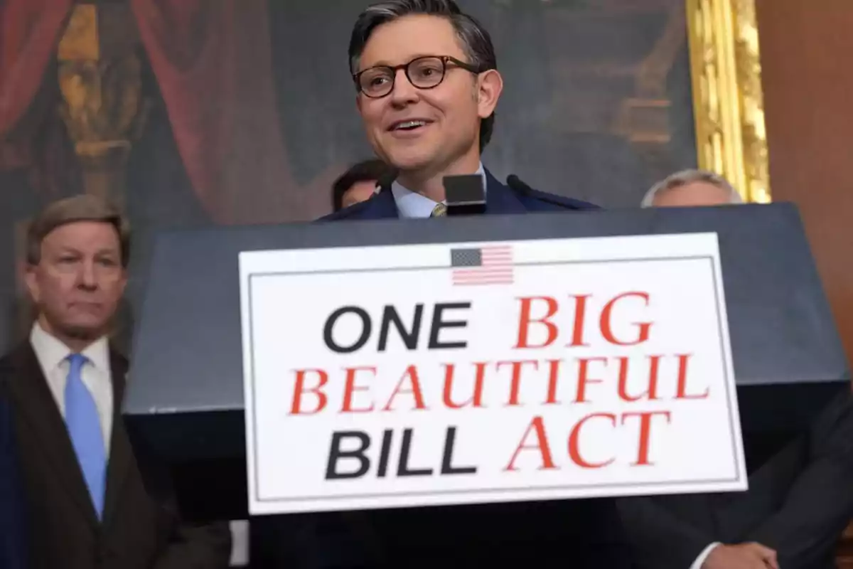 Un hombre con gafas y traje sonríe mientras habla en un podio que tiene un cartel con el texto ONE BIG BEAUTIFUL BILL ACT, acompañado por otras personas al fondo. Un hombre con gafas y traje sonríe mientras habla en un podio que tiene un cartel con el texto ONE BIG BEAUTIFUL BILL ACT, acompañado por otras personas al fondo.