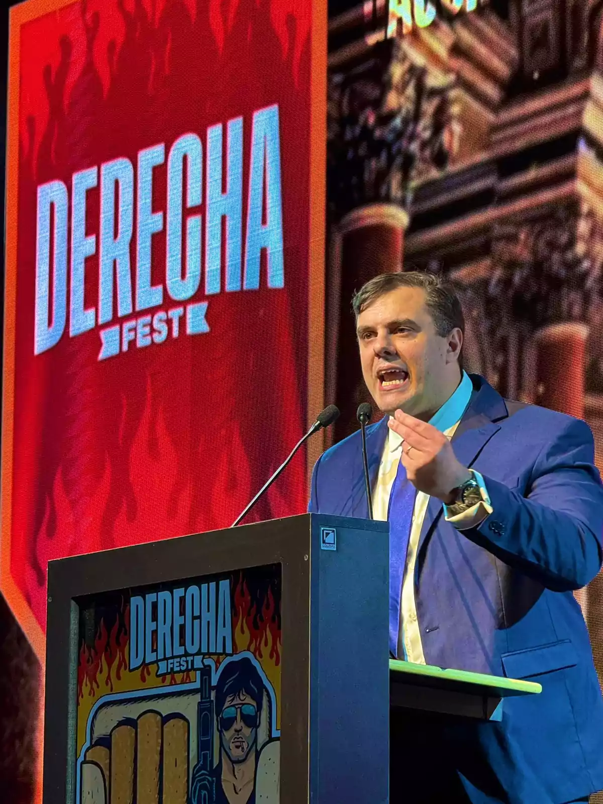 Man in a blue suit speaking at a podium during an event called Derecha Fest, with a red background and a large sign that says Derecha Fest Man in a blue suit speaking at a podium during an event called Derecha Fest, with a red background and a large sign that says Derecha Fest