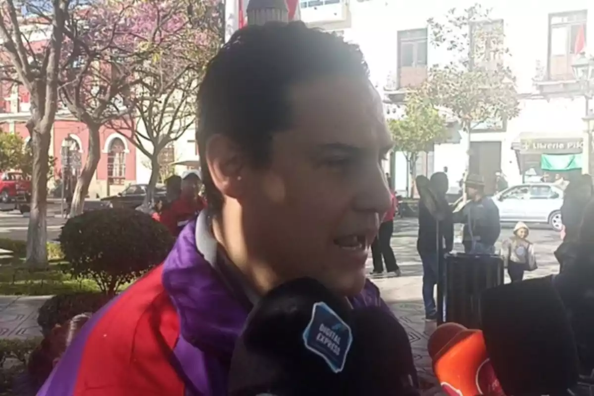 Un hombre con chaqueta roja y morada da una entrevista a varios medios en una plaza pública mientras personas caminan al fondo. Un hombre con chaqueta roja y morada da una entrevista a varios medios en una plaza pública mientras personas caminan al fondo.