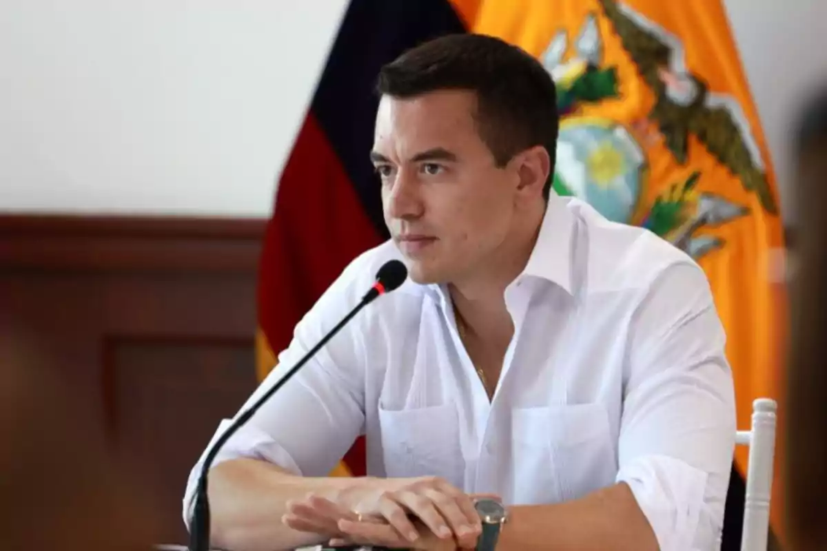 Hombre de cabello corto y camisa blanca hablando frente a un micrófono con una bandera de Ecuador al fondo Hombre de cabello corto y camisa blanca hablando frente a un micrófono con una bandera de Ecuador al fondo