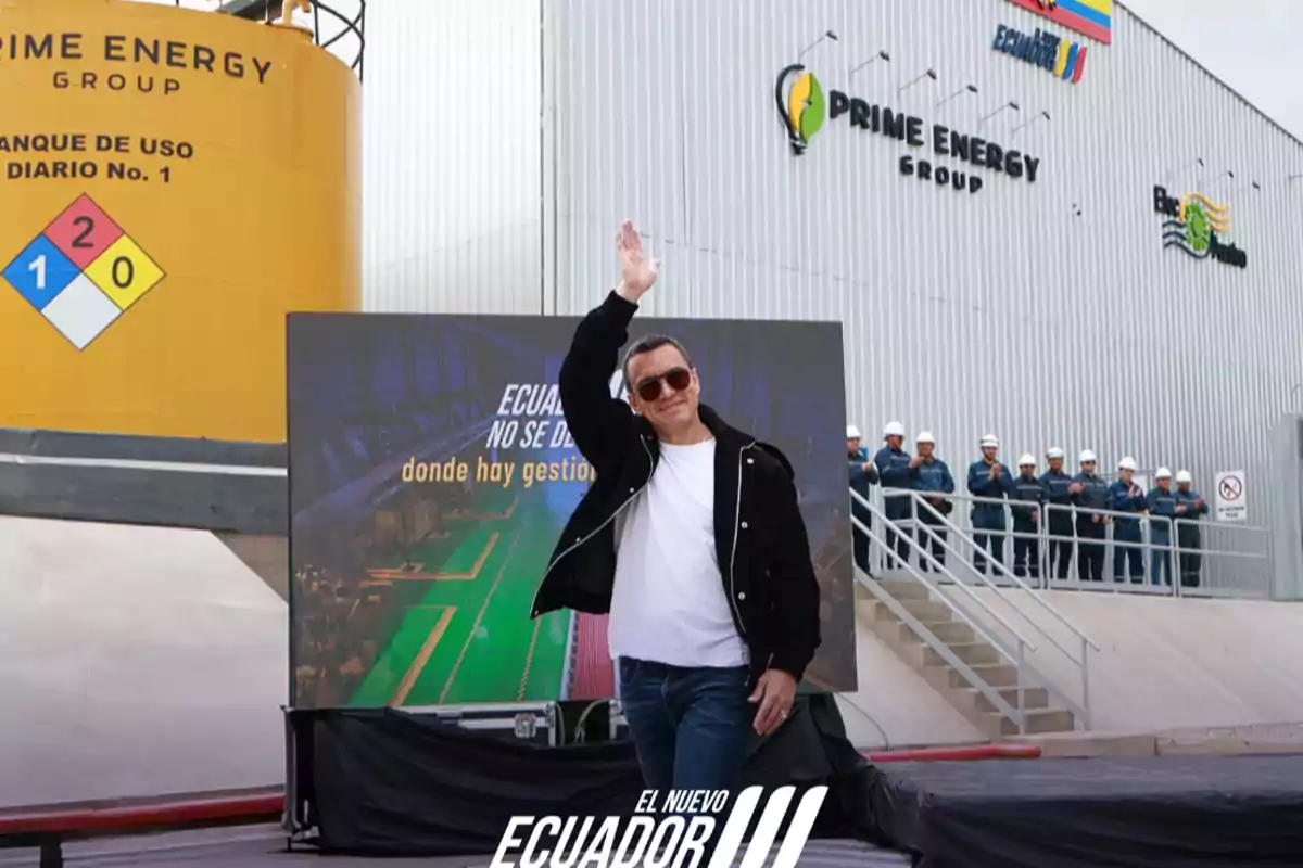Un hombre con gafas de sol y chaqueta negra saluda levantando la mano frente a una planta industrial con el logo de Prime Energy Group y un grupo de trabajadores con casco al fondo