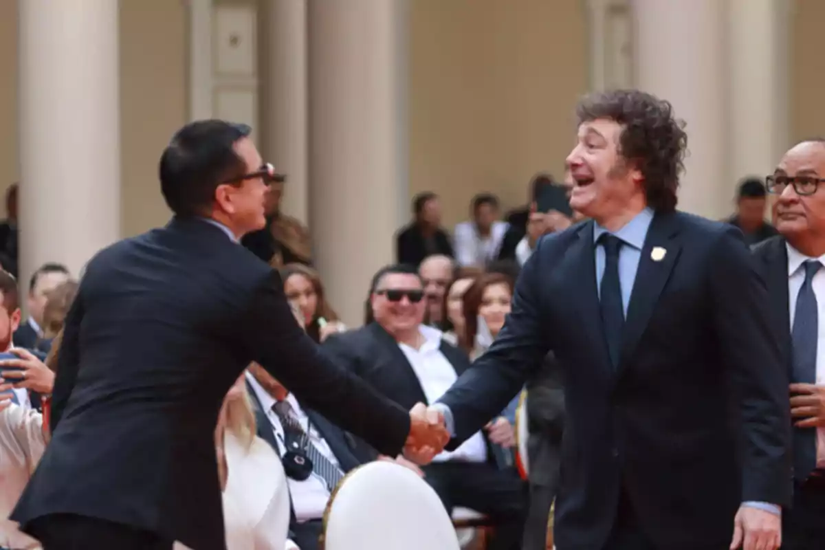 Two men in suits greet each other with a smile and a handshake at a formal event surrounded by other seated people.