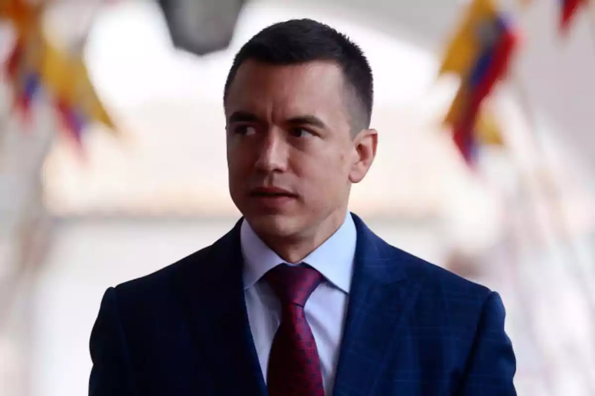 Hombre joven con cabello corto y traje azul con corbata roja mirando hacia un lado con banderas desenfocadas al fondo