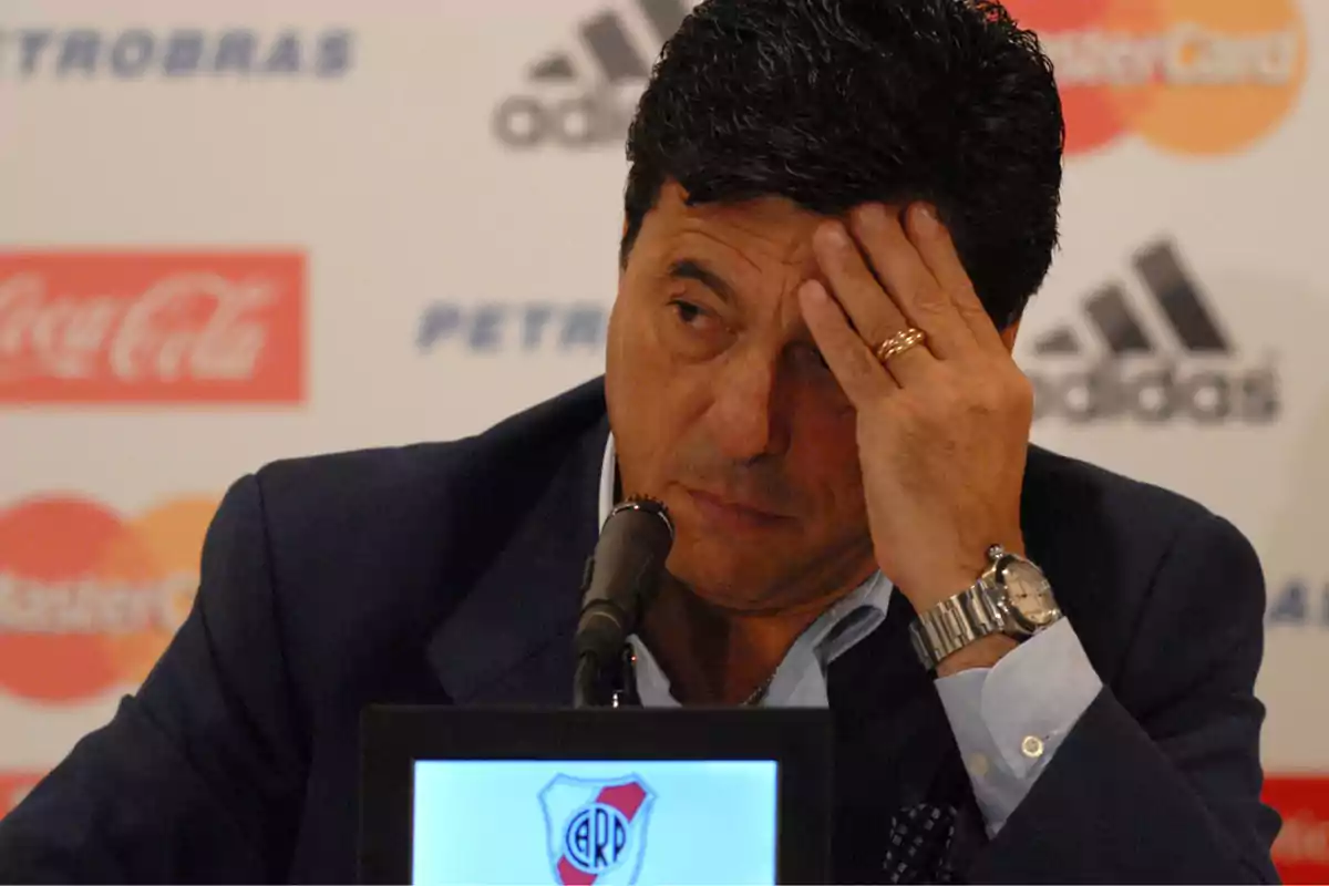 Hombre con expresión preocupada sentado frente a un micrófono en una conferencia de prensa con el escudo de River Plate visible en el podio.