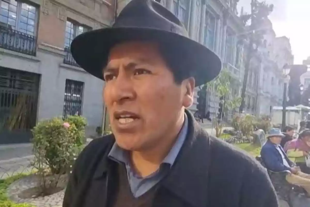 Hombre con sombrero negro hablando en una calle con edificios antiguos y personas sentadas en bancas al fondo