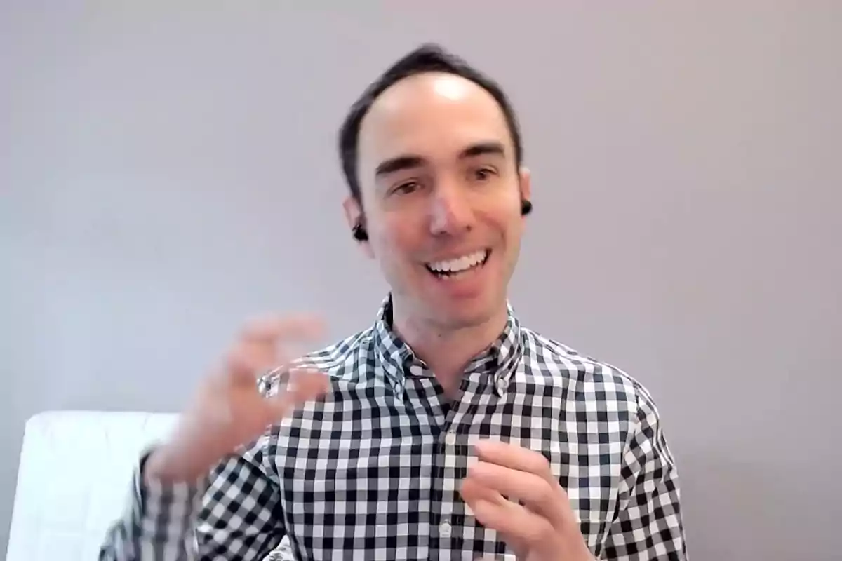 Smiling man in a black and white checkered jersey talking and gesturing in front of a gray wall Smiling man in a black and white checkered jersey talking and gesturing in front of a gray wall