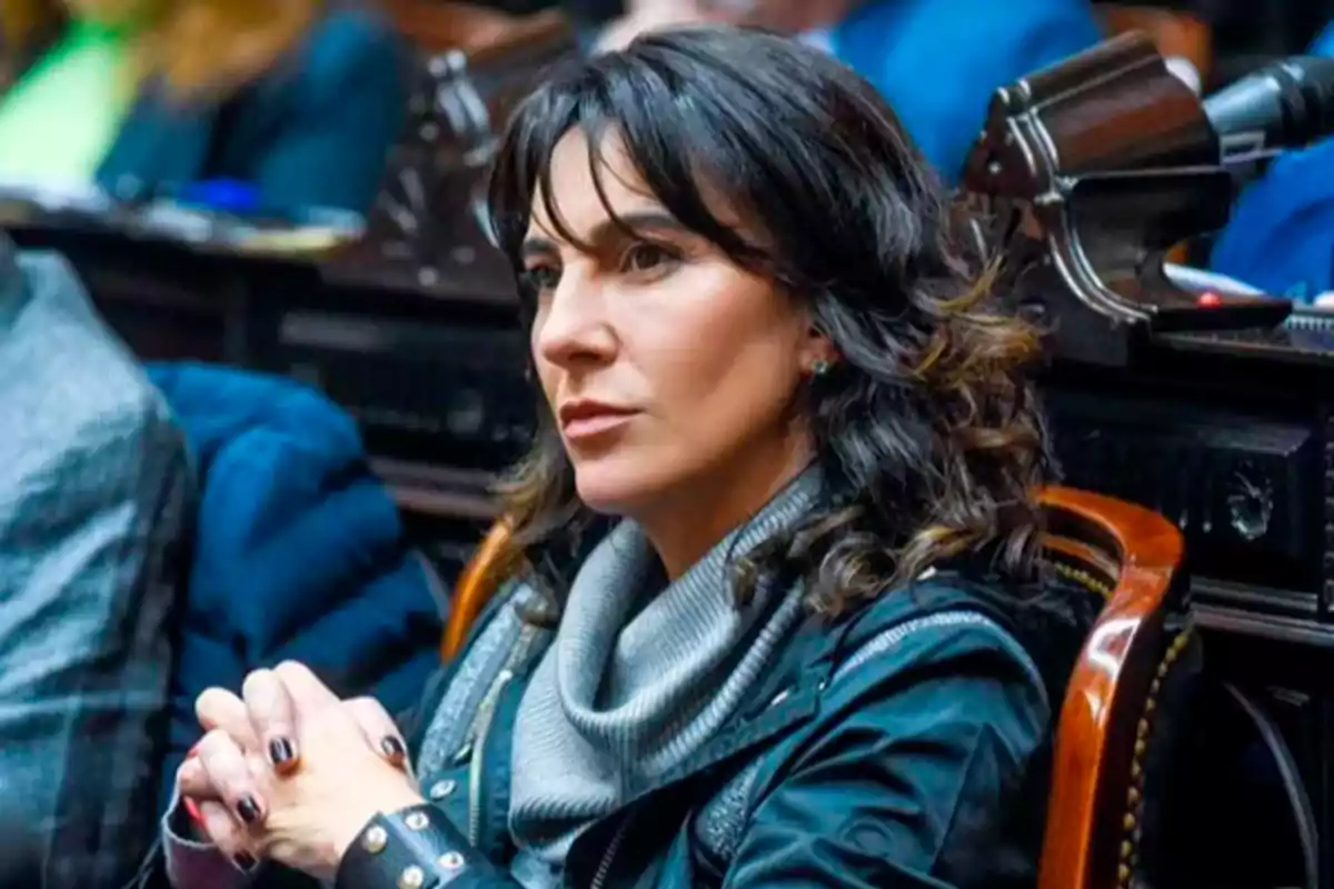 Mujer de cabello oscuro sentada en una silla de madera con expresión seria y las manos entrelazadas Mujer de cabello oscuro sentada en una silla de madera con expresión seria y las manos entrelazadas