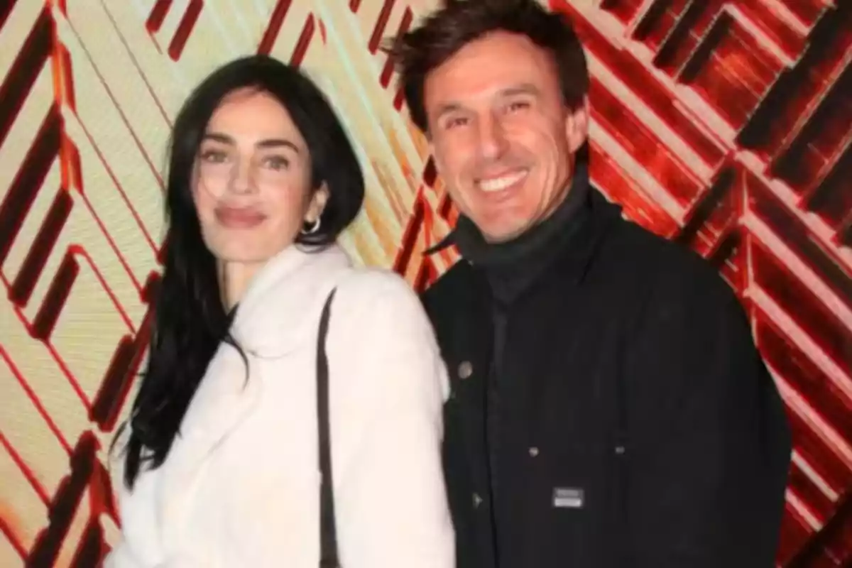 A couple smiles in front of a background with geometric patterns in shades of red and beige. A couple smiles in front of a background with geometric patterns in shades of red and beige.