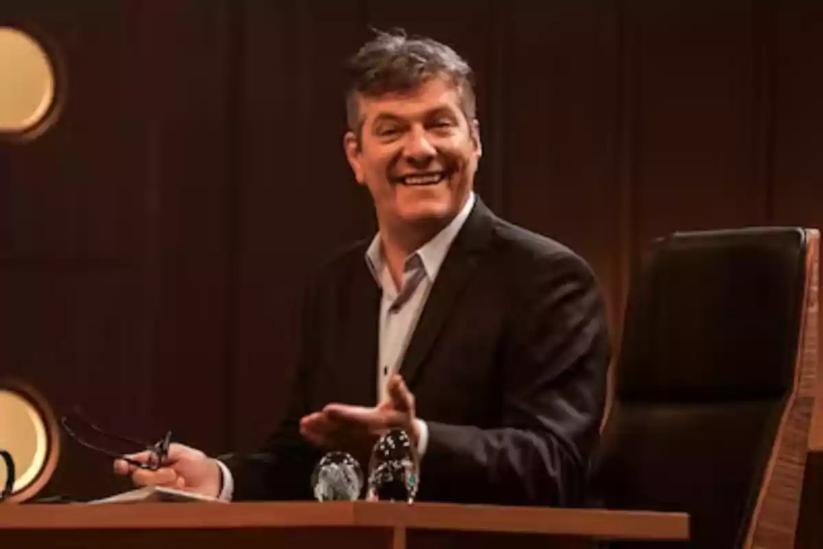 Hombre de traje oscuro sonriendo mientras está sentado en un escritorio de madera en un ambiente elegante Hombre de traje oscuro sonriendo mientras está sentado en un escritorio de madera en un ambiente elegante