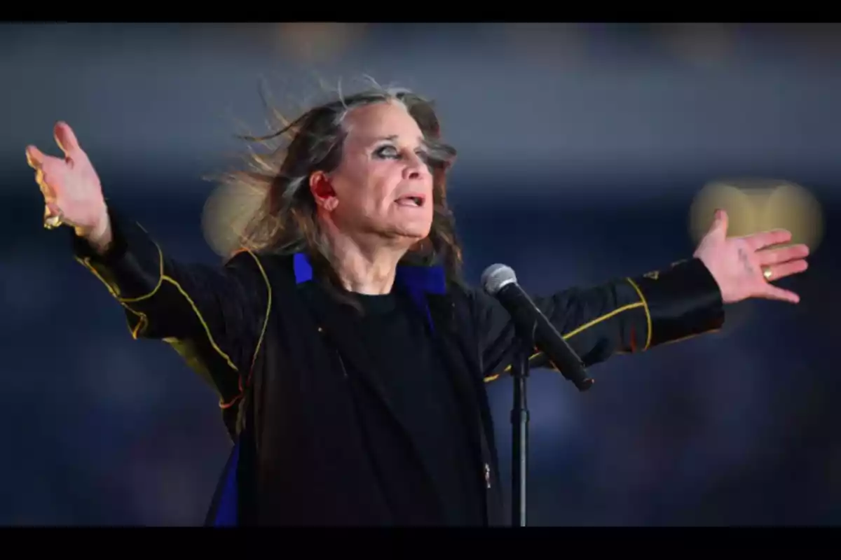 Persona con el cabello largo y maquillaje oscuro cantando en un escenario con los brazos extendidos frente a un micrófono Persona con el cabello largo y maquillaje oscuro cantando en un escenario con los brazos extendidos frente a un micrófono