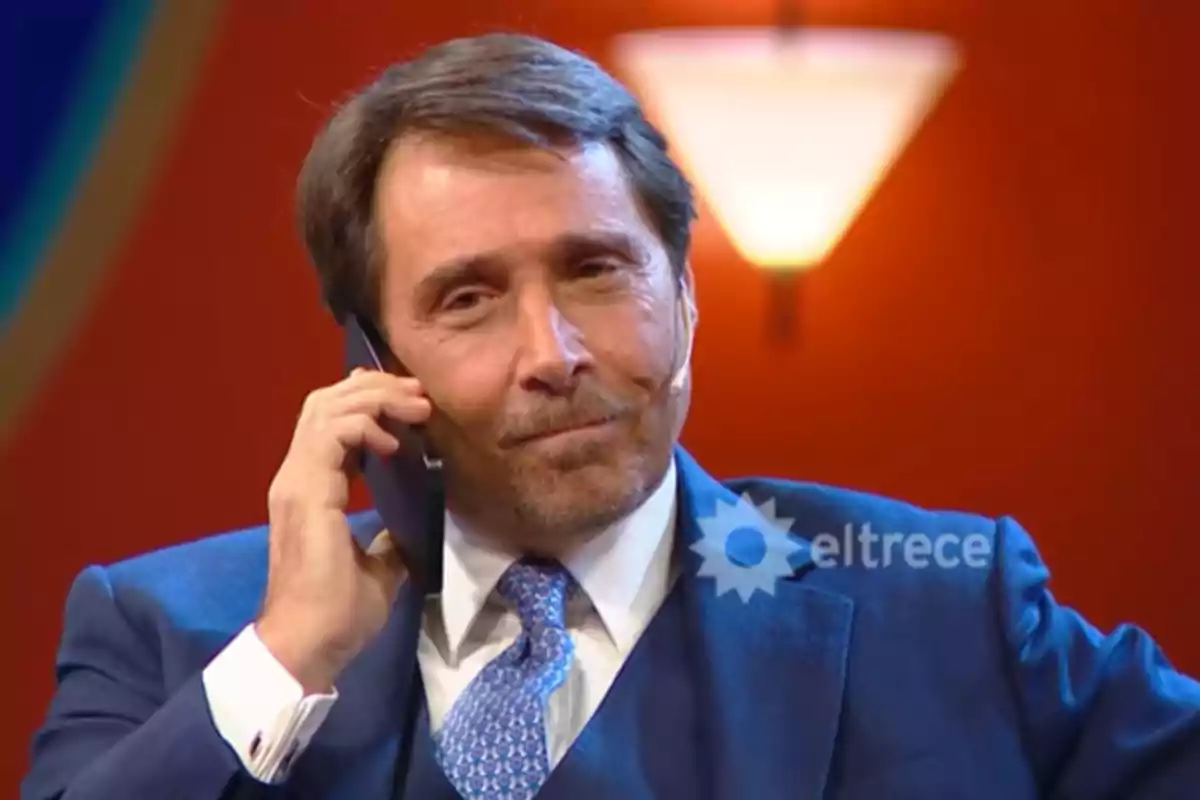 Hombre de traje azul hablando por teléfono en un set de televisión con fondo naranja y logo de eltrece Hombre de traje azul hablando por teléfono en un set de televisión con fondo naranja y logo de eltrece