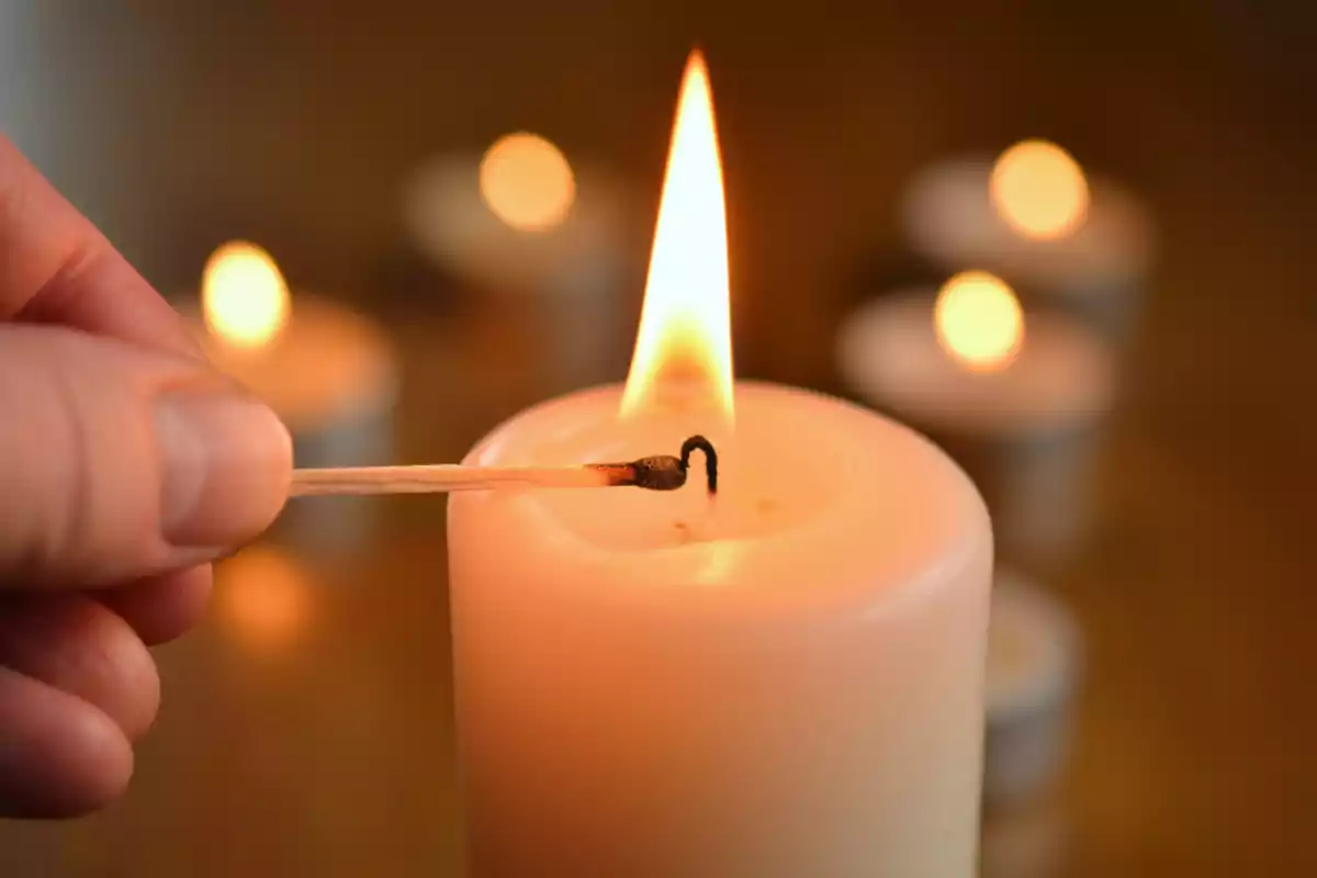 Mano encendiendo una vela blanca con un fósforo mientras varias velas desenfocadas están al fondo
