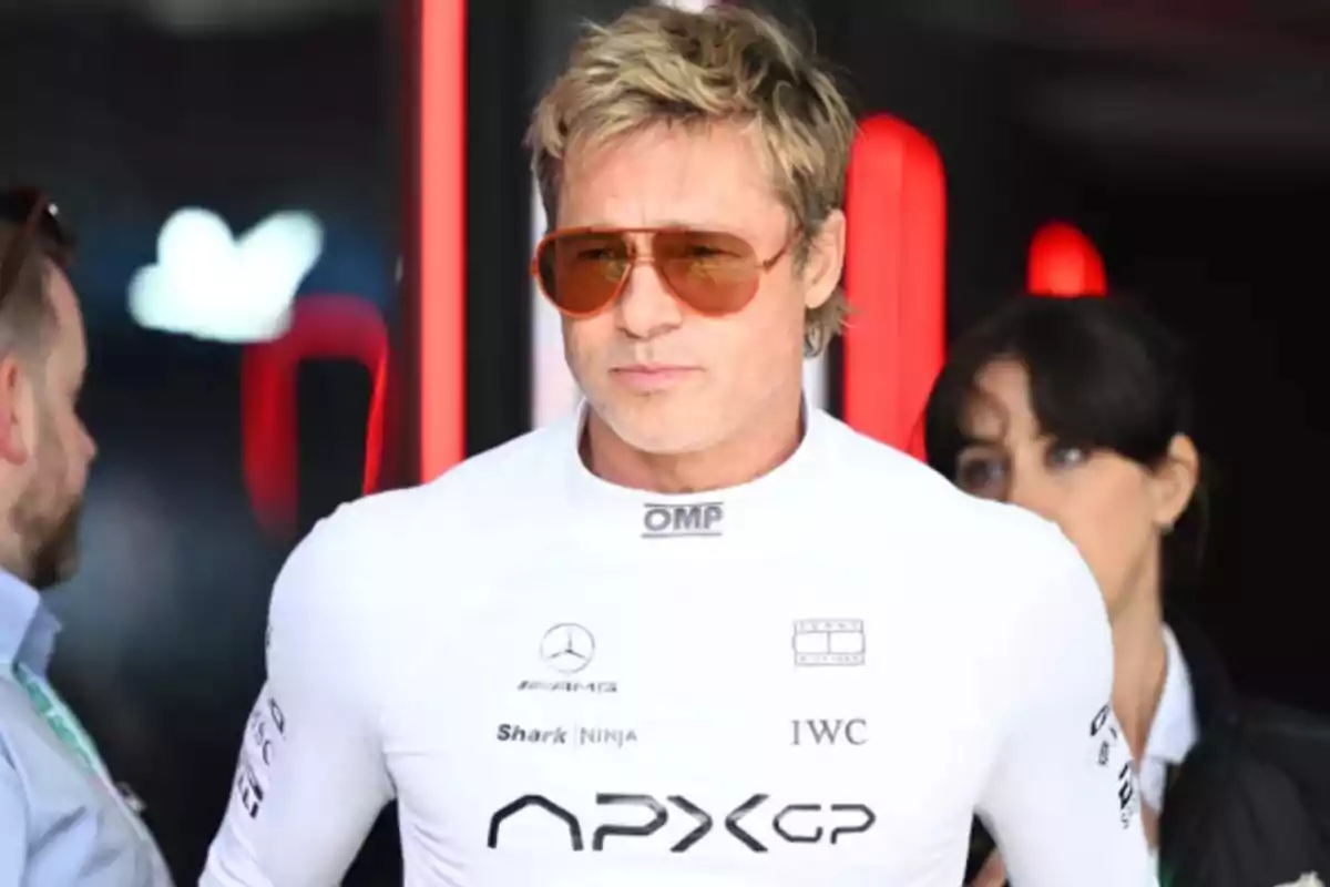 Blond man wearing sunglasses and a white racing driver jersey with several logos, in a motorsport competition setting.