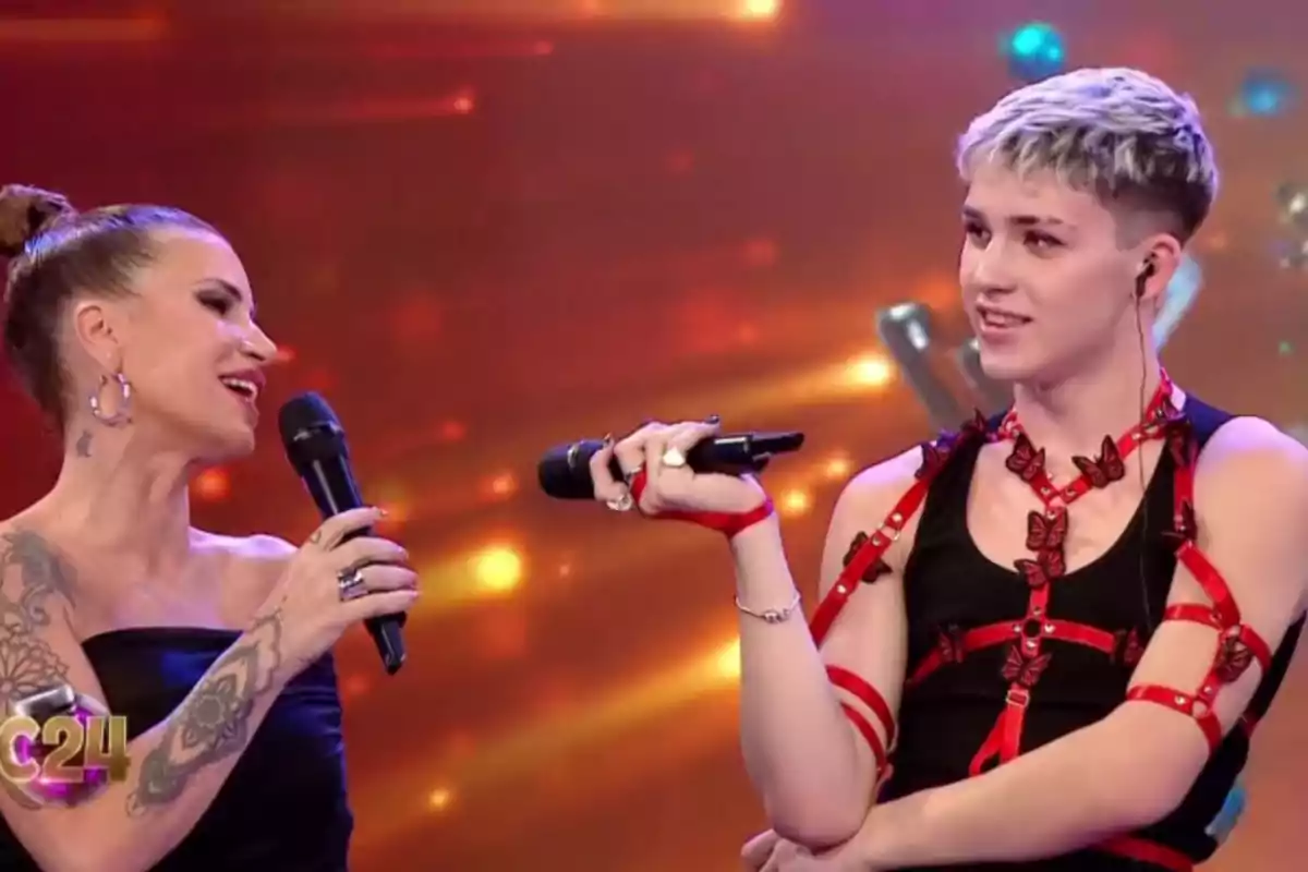 Two people on a television stage holding microphones and talking, one of them wearing a black outfit with red stripes and decorative butterflies. Two people on a television stage holding microphones and talking, one of them wearing a black outfit with red stripes and decorative butterflies.