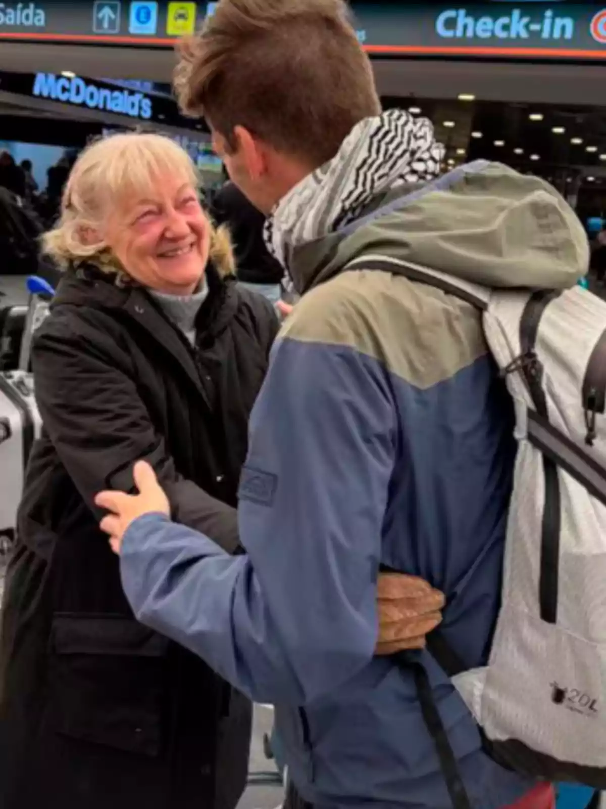 An older woman smiles as she hugs a young man with a backpack at what appears to be the entrance of an airport. An older woman smiles as she hugs a young man with a backpack at what appears to be the entrance of an airport.