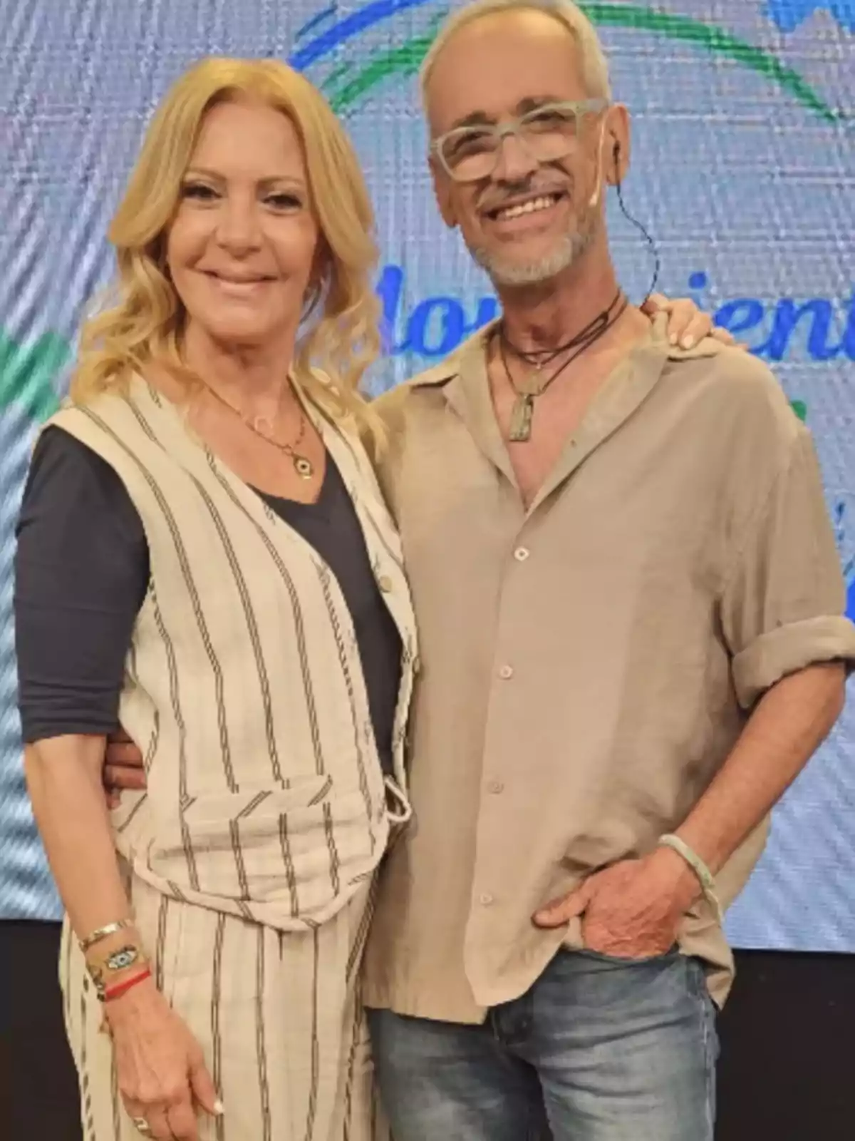 Two adults smile as they pose in front of a colorful screen, a blonde woman wearing a vest and striped pants and a gray-haired man with glasses and a beige jersey. Two adults smile as they pose in front of a colorful screen, a blonde woman wearing a vest and striped pants and a gray-haired man with glasses and a beige jersey.