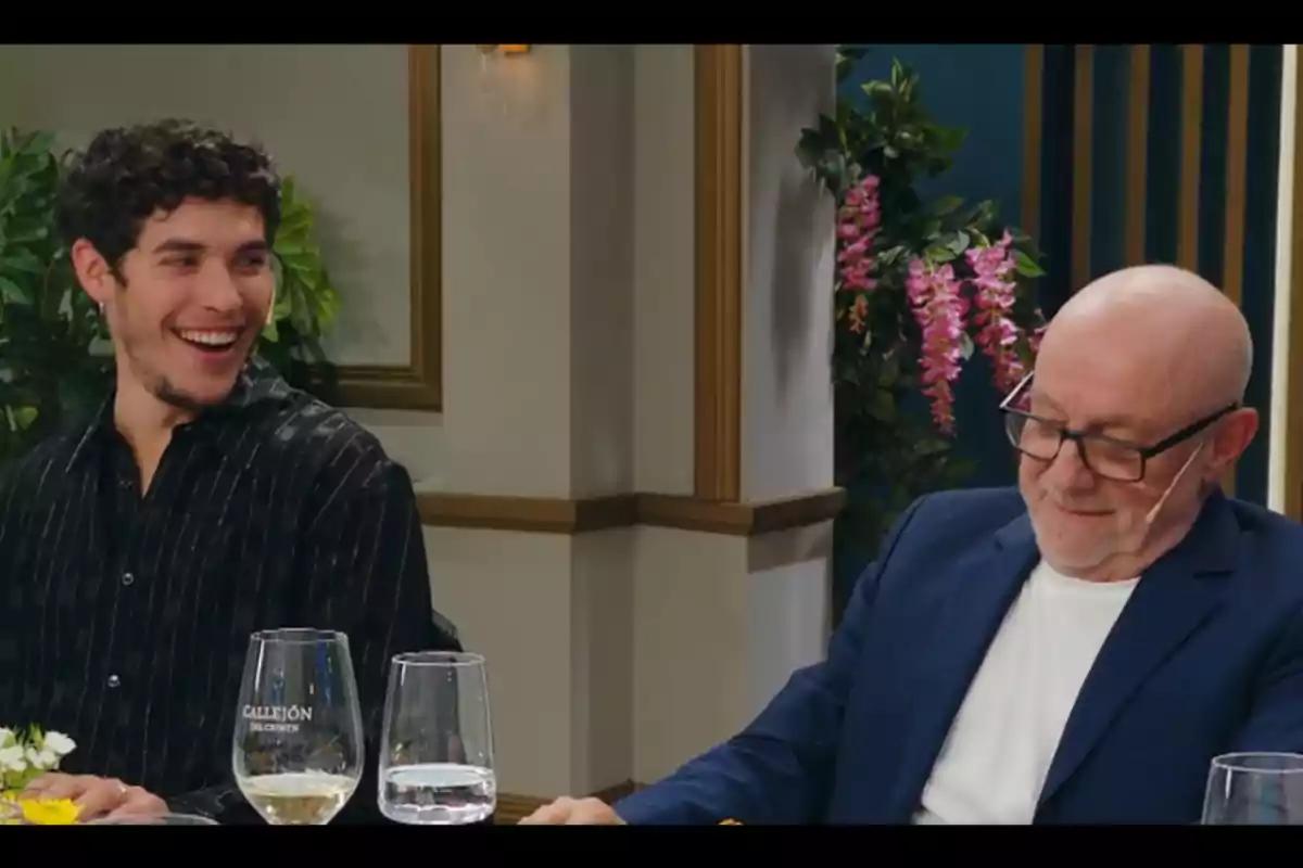 Two men are sitting at an elegant table; one of them is smiling while the other looks down with glasses and a relaxed expression. In the background, there are decorative plants and wine glasses on the table. Two men are sitting at an elegant table; one of them is smiling while the other looks down with glasses and a relaxed expression. In the background, there are decorative plants and wine glasses on the table.