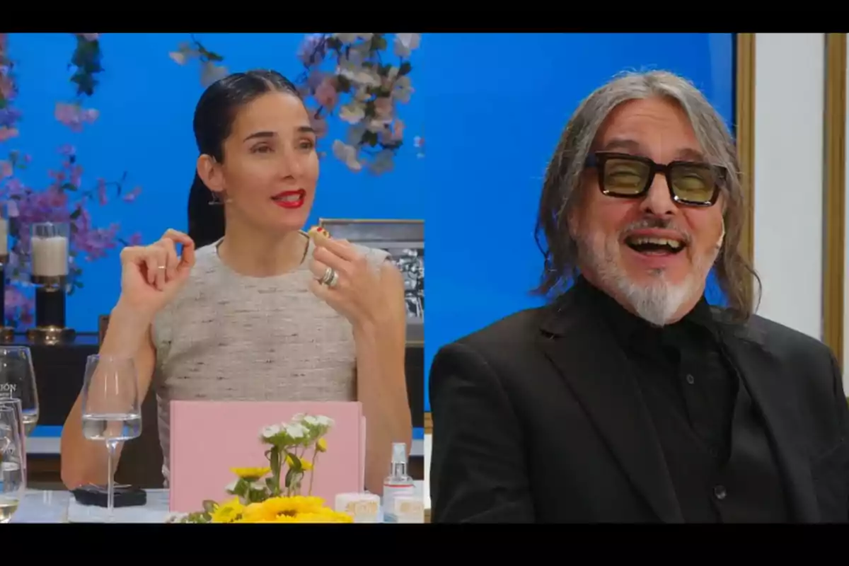 A dark-haired woman and a man with glasses and a gray beard smile as they talk at a table decorated with flowers and glasses, with a blue background and pink floral details. A dark-haired woman and a man with glasses and a gray beard smile as they talk at a table decorated with flowers and glasses, with a blue background and pink floral details.