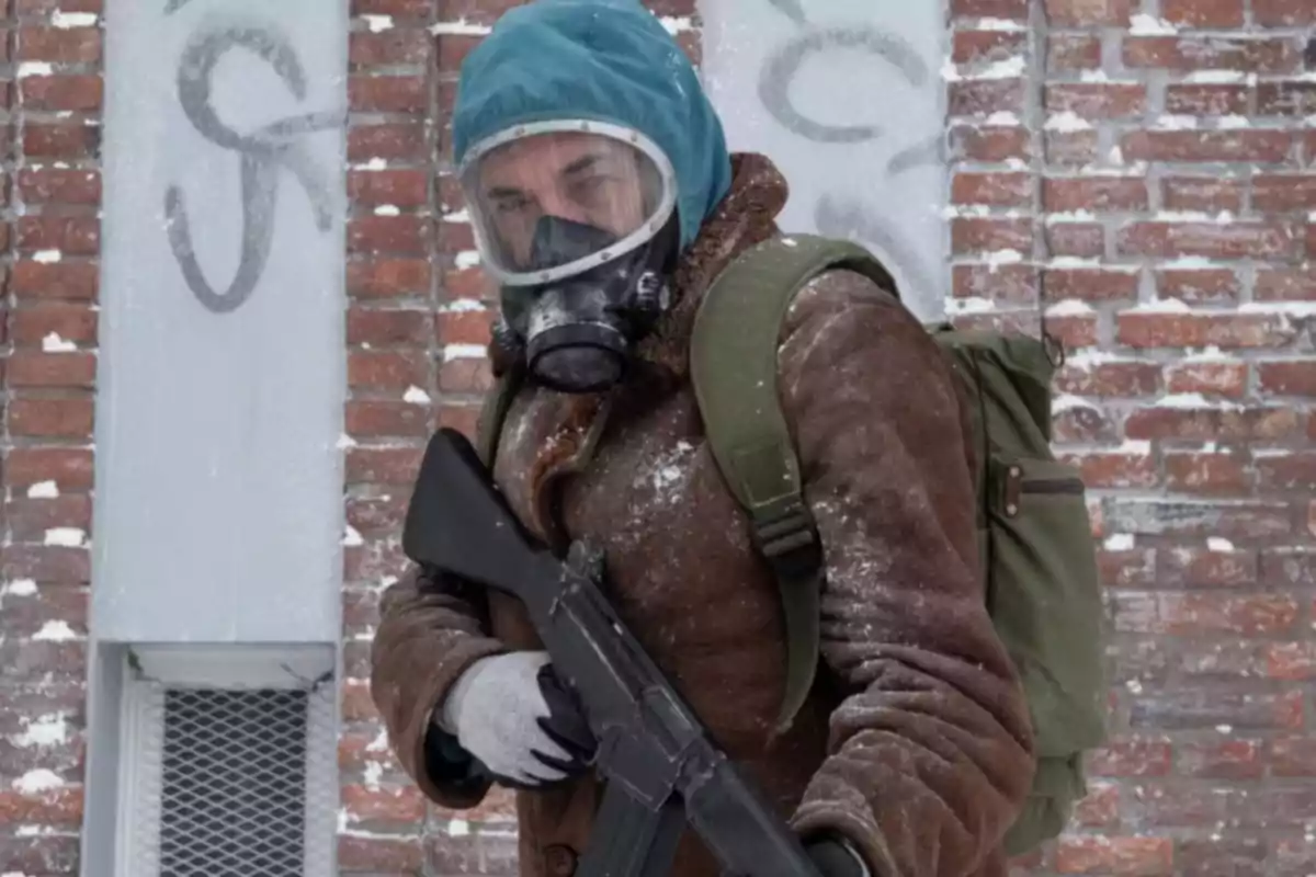 Person wearing a brown coat, green backpack, and gas mask holding a weapon in a snowy environment in front of a brick wall