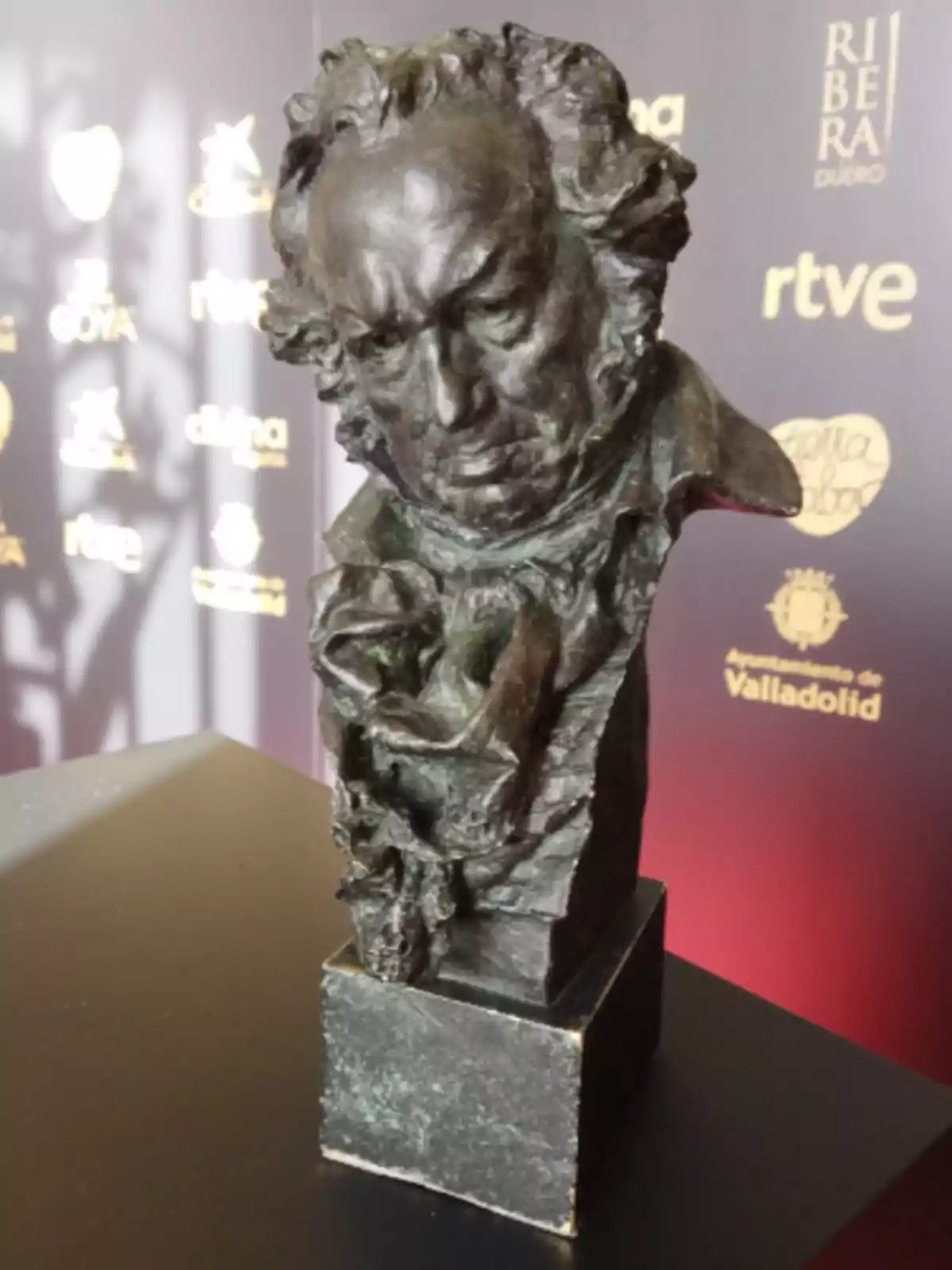 Escultura de un busto de bronce sobre una base cuadrada, con fondo de logotipos y letras doradas desenfocadas.