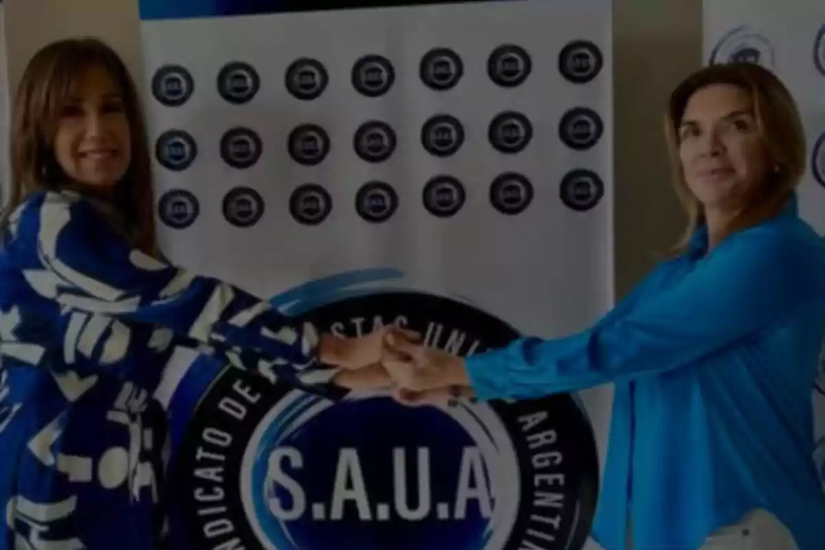 Dos mujeres sonríen y se dan la mano frente a un cartel del Sindicato de Artistas Unidos Argentinos Dos mujeres sonríen y se dan la mano frente a un cartel del Sindicato de Artistas Unidos Argentinos