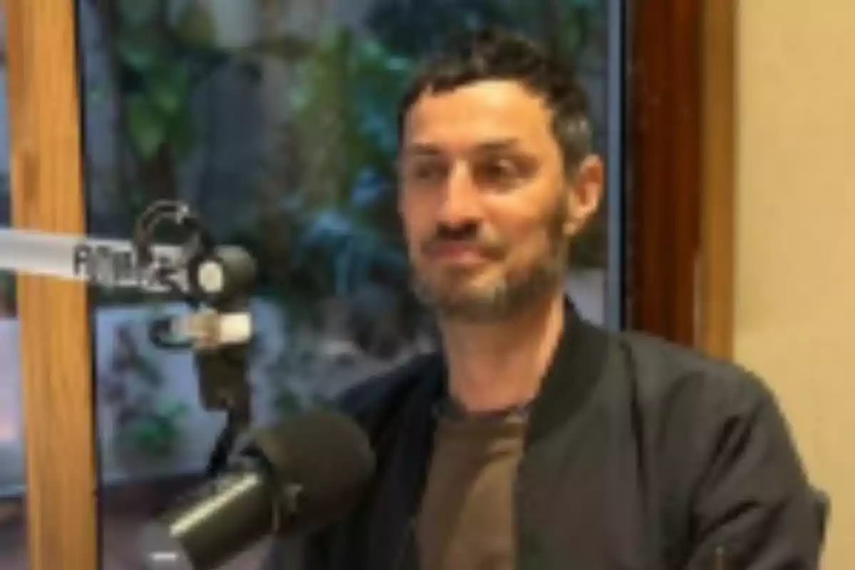 Hombre de cabello corto y barba sentado frente a un micrófono en un estudio de grabación con ventana al fondo Hombre de cabello corto y barba sentado frente a un micrófono en un estudio de grabación con ventana al fondo