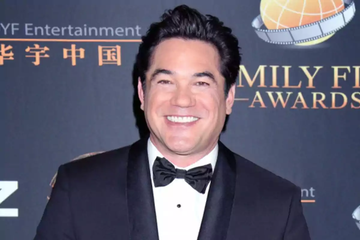 Dark-haired man in a tuxedo and black bow tie smiling on a red carpet in front of a background with golden letters. Dark-haired man in a tuxedo and black bow tie smiling on a red carpet in front of a background with golden letters.