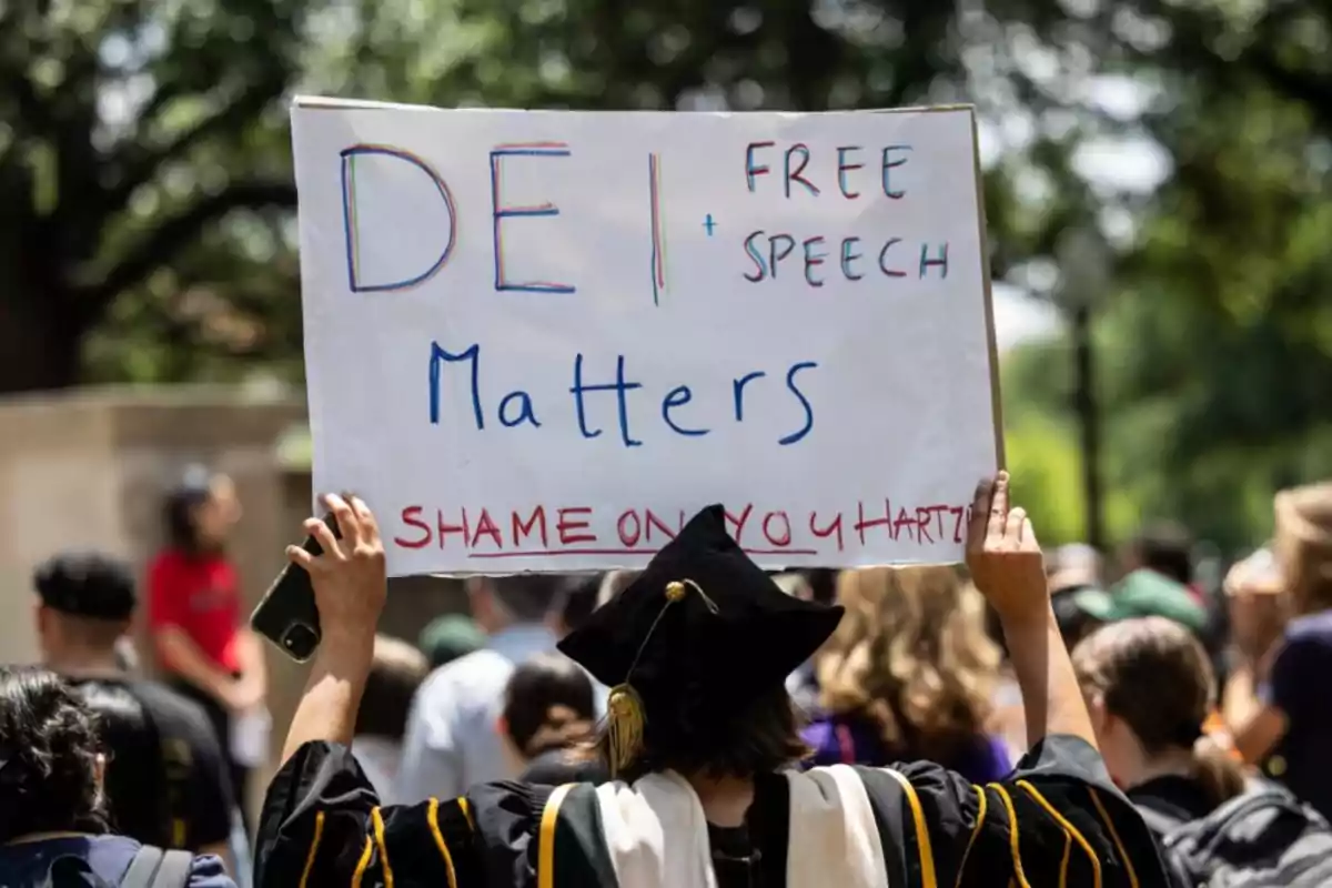 Person in cap and gown holds a sign at a university protest that says DEI Free Speech Matters Shame on you Hartzell while other people are in the background Person in cap and gown holds a sign at a university protest that says DEI Free Speech Matters Shame on you Hartzell while other people are in the background