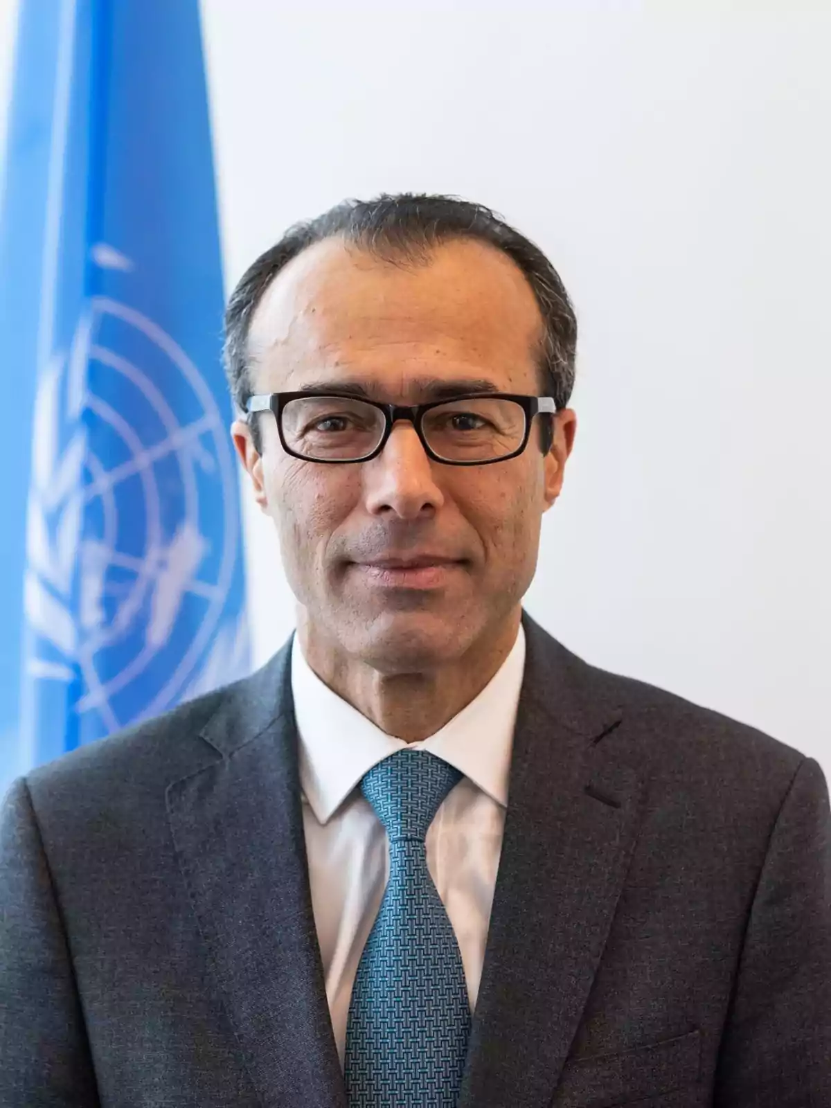 Hombre de mediana edad con gafas y traje formal posando frente a una bandera azul con el logotipo de la ONU Hombre de mediana edad con gafas y traje formal posando frente a una bandera azul con el logotipo de la ONU