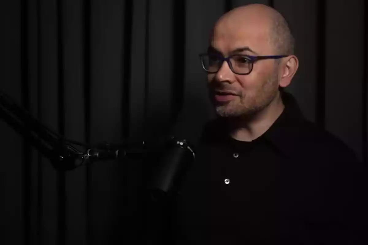 Hombre calvo con gafas y barba corta hablando frente a un micrófono en un estudio con cortinas oscuras de fondo Hombre calvo con gafas y barba corta hablando frente a un micrófono en un estudio con cortinas oscuras de fondo