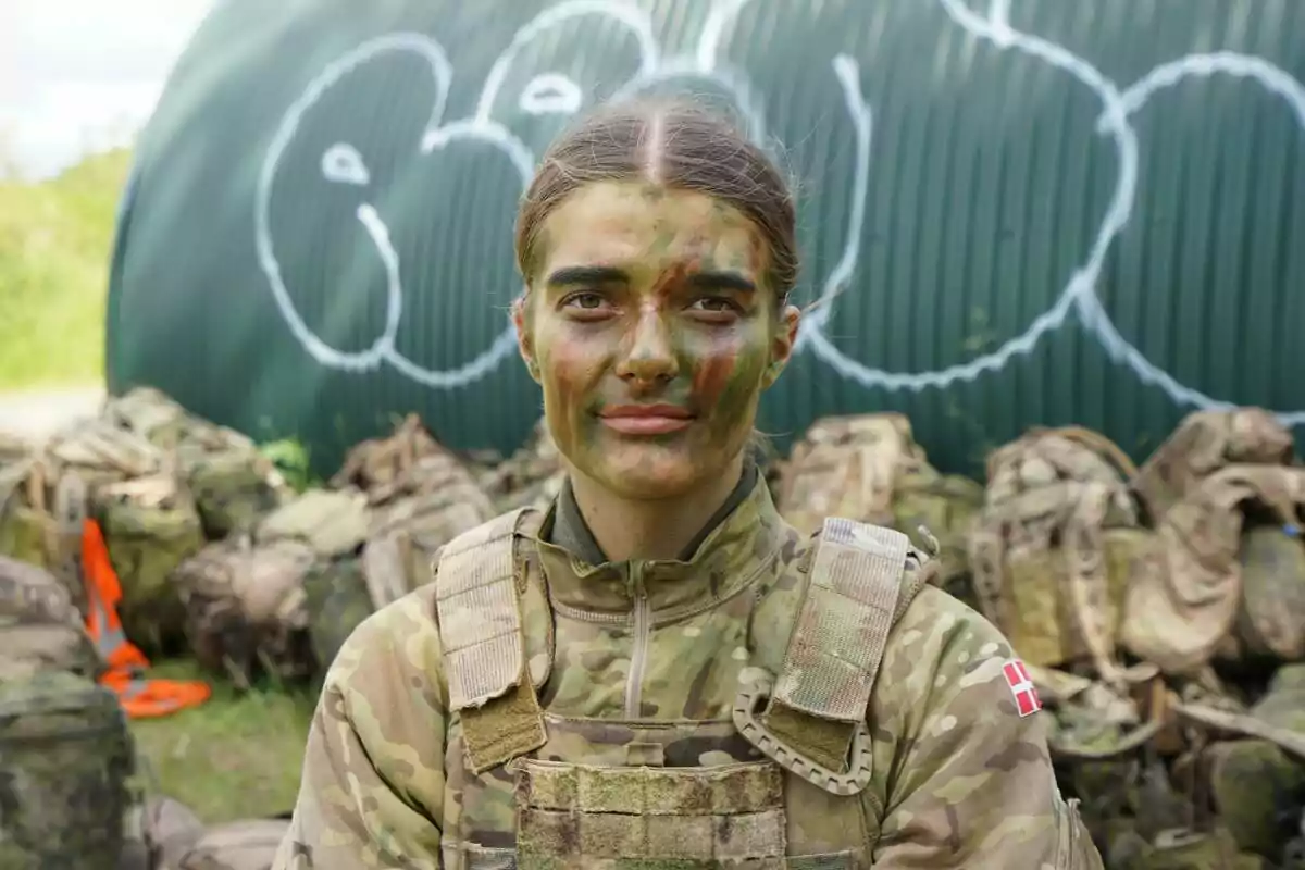 Mujer soldado con uniforme de camuflaje y pintura en el rostro, de pie frente a mochilas militares y una estructura verde con grafiti blanco