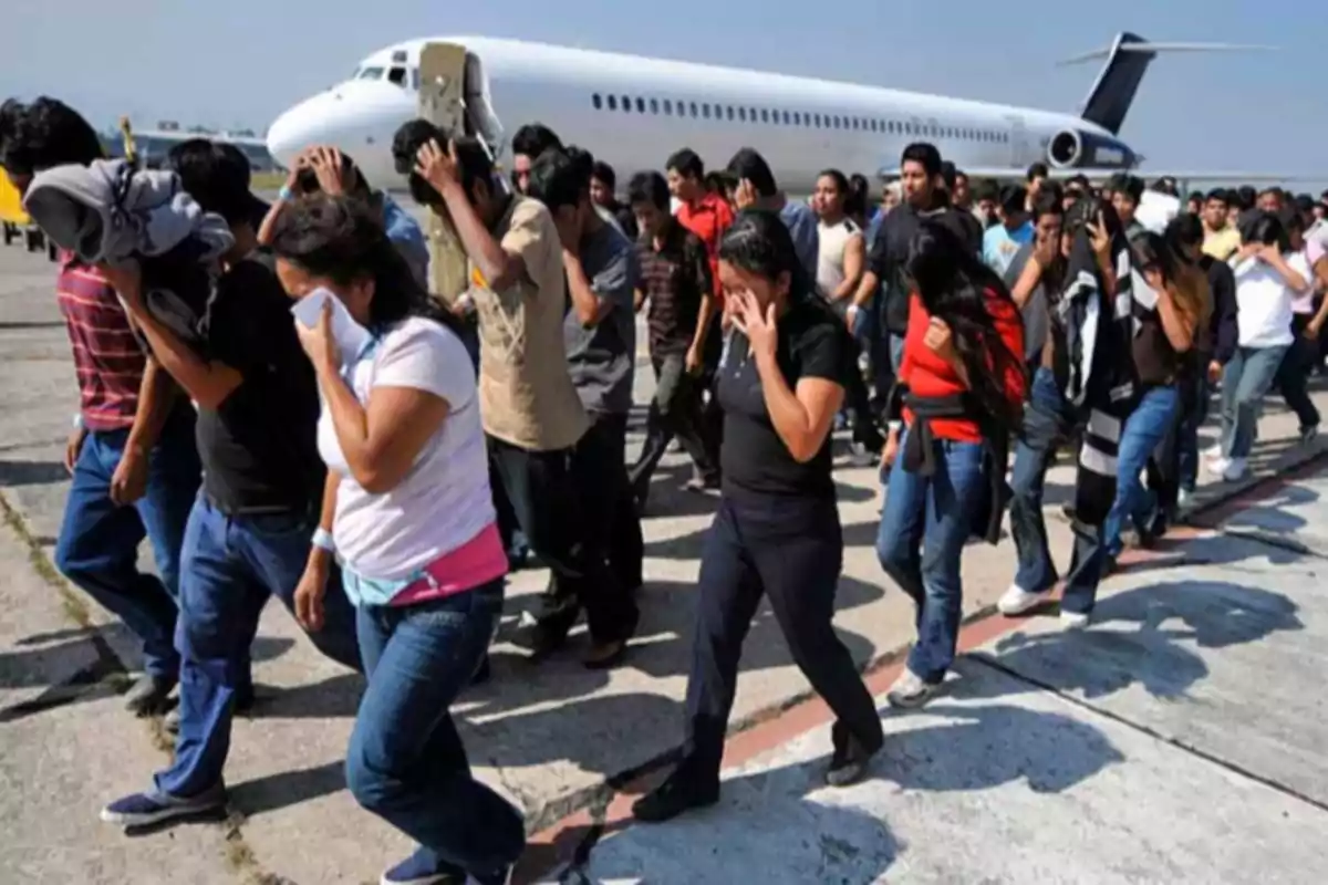 Un grupo numeroso de personas camina en fila junto a un avión en una pista, muchas de ellas cubriéndose el rostro con las manos o ropa.