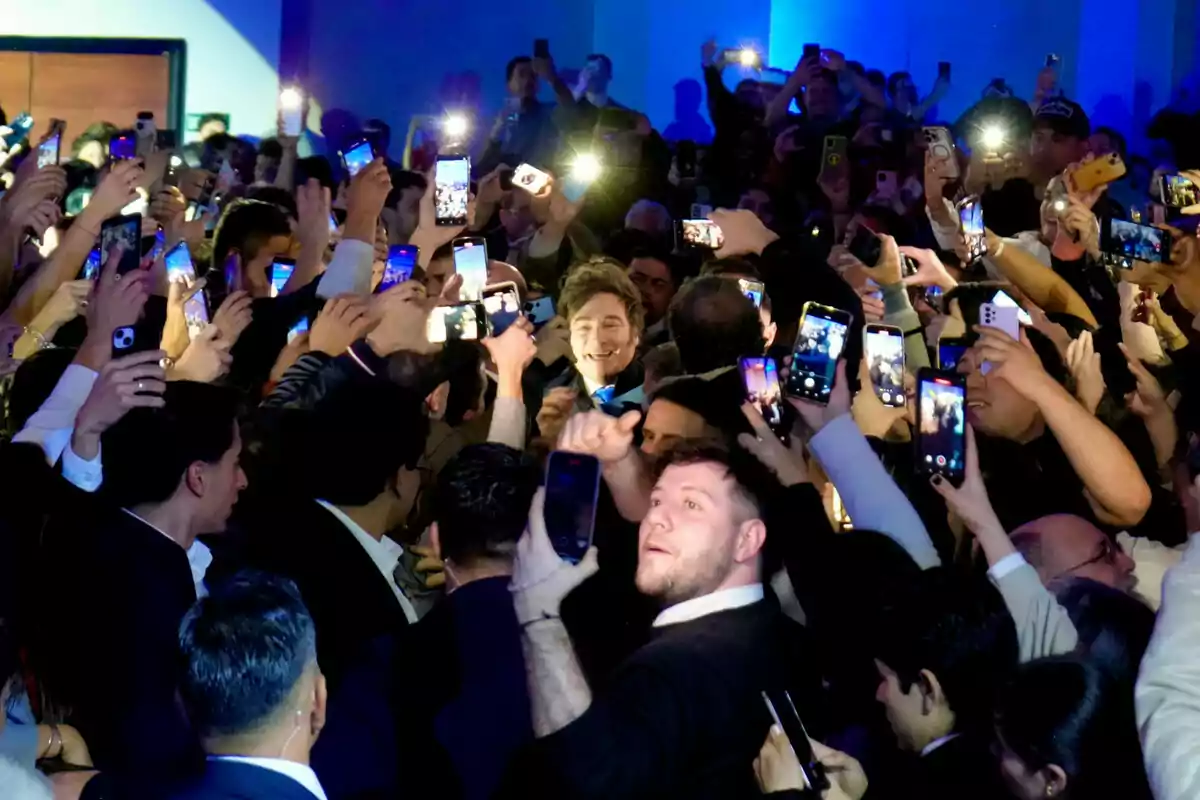 Un hombre sonriente rodeado de una multitud que lo fotografía con sus teléfonos móviles en un ambiente iluminado por flashes y pantallas. Un hombre sonriente rodeado de una multitud que lo fotografía con sus teléfonos móviles en un ambiente iluminado por flashes y pantallas.