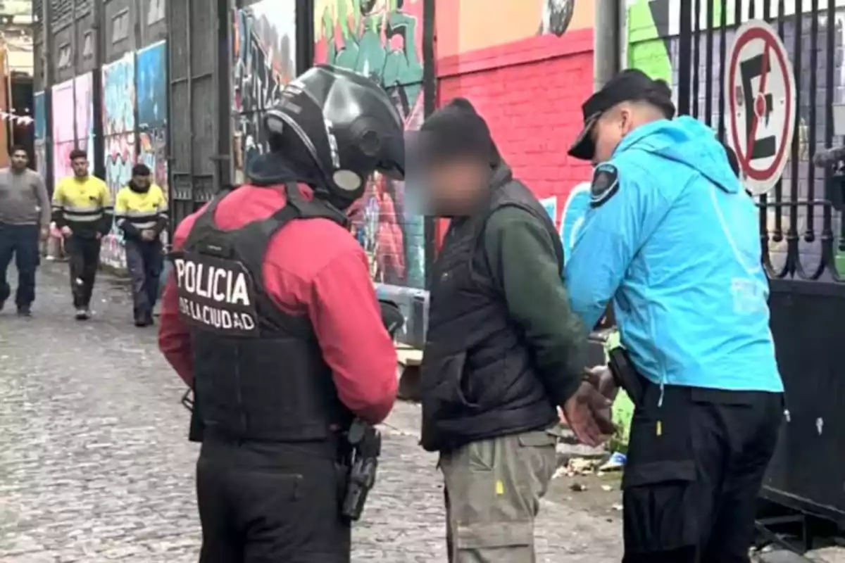 Policías detienen a un hombre en una calle con murales de colores mientras otras personas caminan al fondo Policías detienen a un hombre en una calle con murales de colores mientras otras personas caminan al fondo
