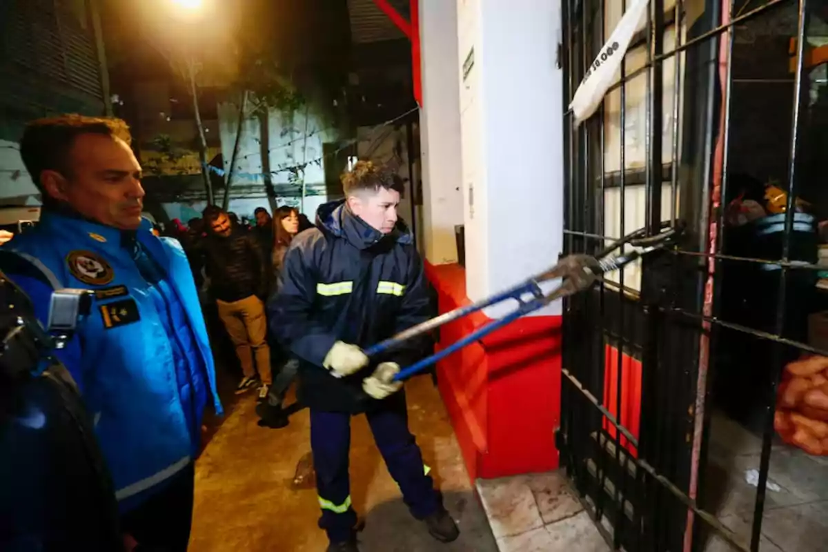Un bombero utiliza una cizalla para cortar los barrotes de una puerta de metal mientras varias personas observan la escena en la calle durante la noche